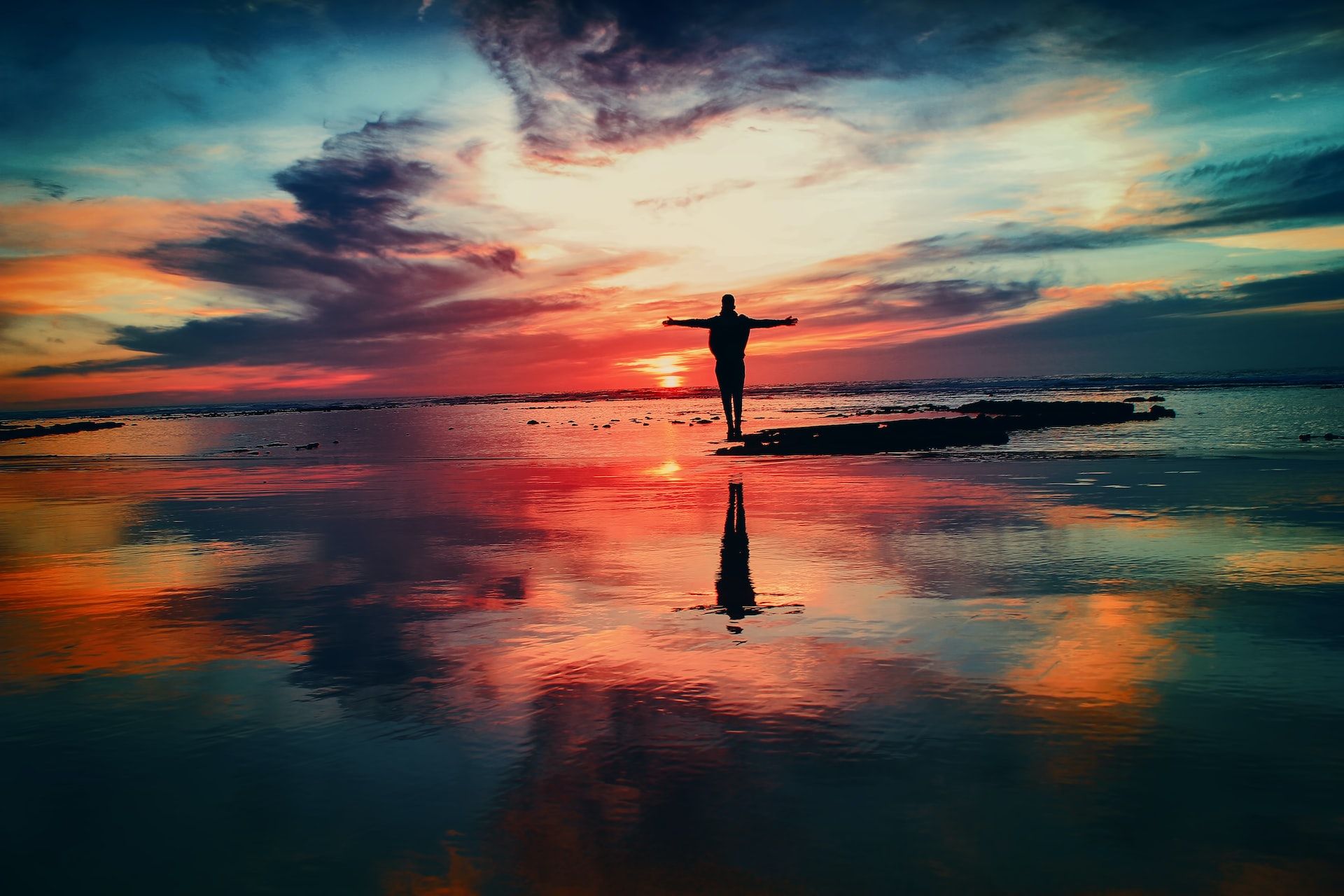 Eine Person steht mit ausgebreiteten Armen am Strand vor einem wolkenverhangenen rotem Sonnenuntergang.